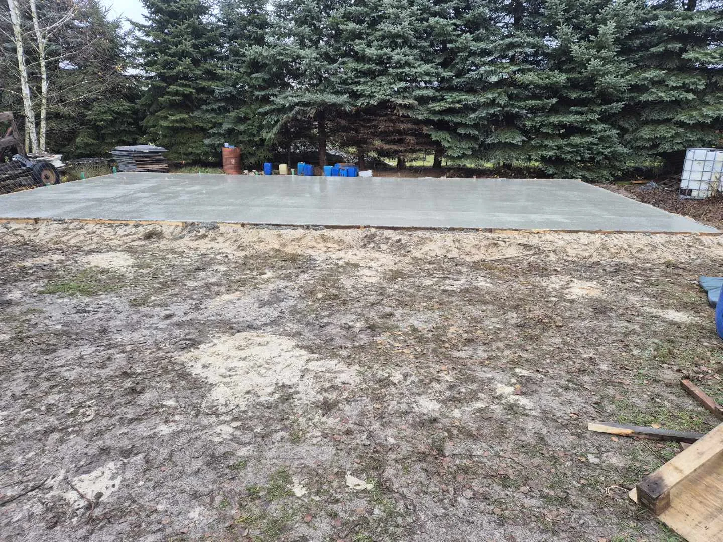 Na zdjęciu prezentuje się świeżo utwardzona posadzka betonowa na podbudowie, otoczona terenem budowlanym i drzewostanem