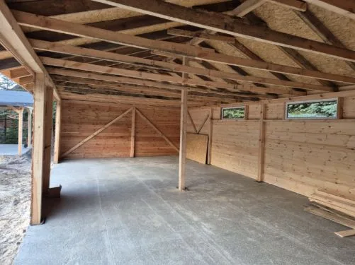 Wnętrze drewnianego budynku gospodarczego w trakcie budowy, z widoczną konstrukcją stropu i surowymi ścianami